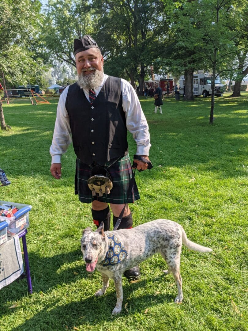 A man in a kilt and dog are standing on the grass.